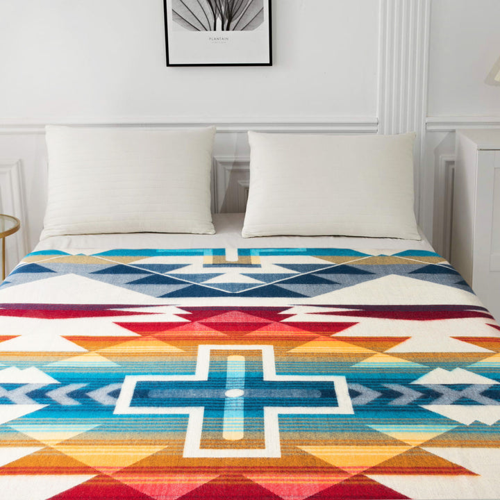 Bedspread displays colorful geometric patterns with a central cross design, draped over a neatly made bed with two pillows. A minimalist artwork labeled PLANTAIN hangs above in a subtle room.