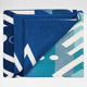 A folded blue beach towel features geometric white and turquoise patterns, with overlapping layers. It rests on a plain, light gray background.