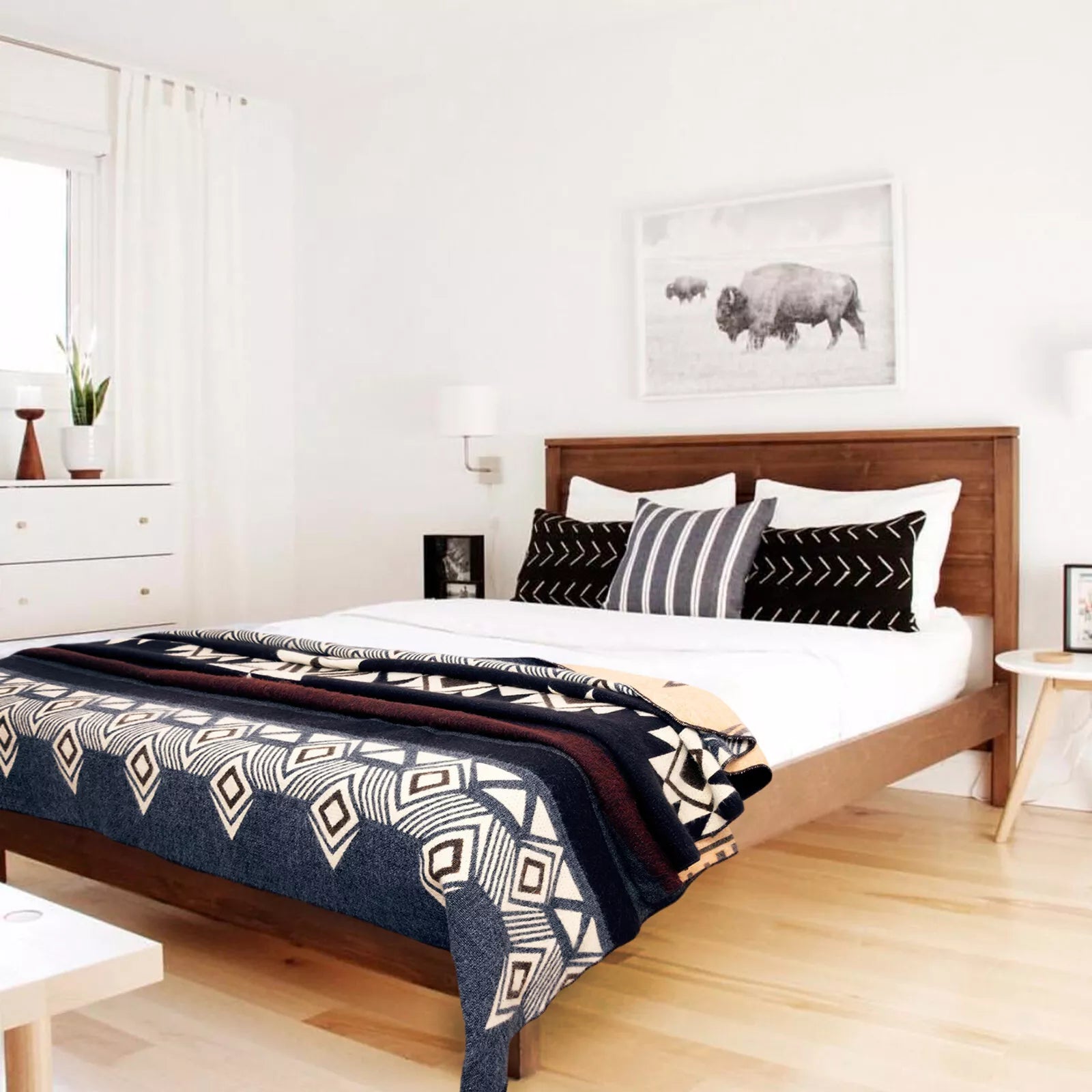 A neatly made bed, featuring patterned pillows and a geometric blanket, sits in a bright room with wooden floors. A dresser with plants and a bison artwork decorate the space.