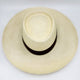 A light cream-colored, wide-brimmed straw hat with a dark brown band lies on a neutral surface, showcasing its woven texture and round crown design.