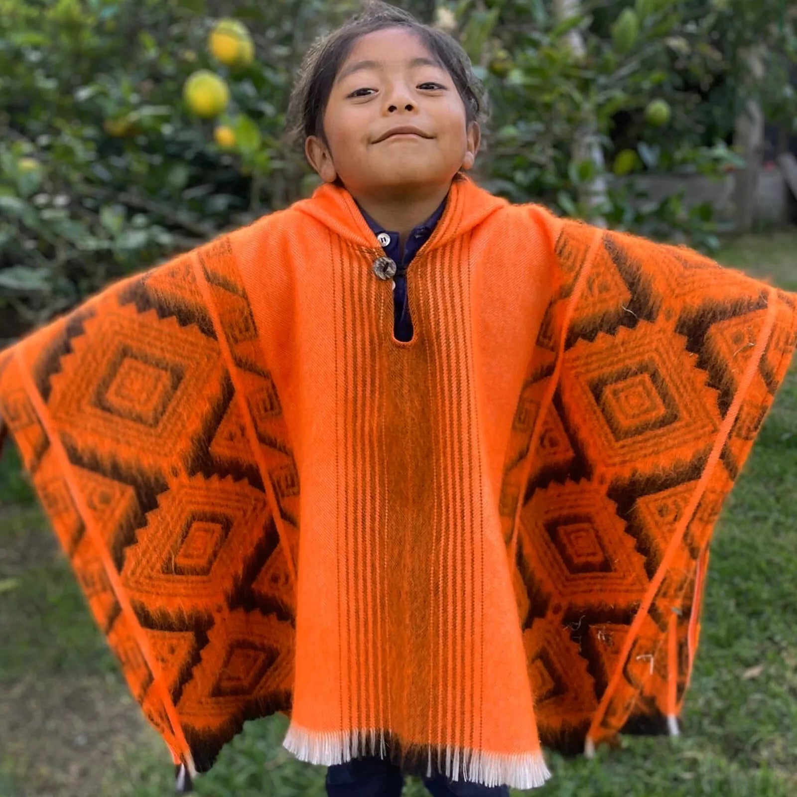 A child wearing a vibrant orange poncho with geometric patterns stands confidently outdoors, surrounded by green foliage and small fruit trees.