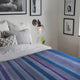 Morona baby alpaca wool blanket – a bed with gray and patterned pillows is covered by a striped blanket. A wall-mounted lamp emits light beside framed artwork and a vase with yellow flowers rests on a table