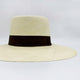 A wide-brimmed straw hat with a dark brown ribbon rests in a neutral, well-lit setting, highlighting its texture and elegant design.