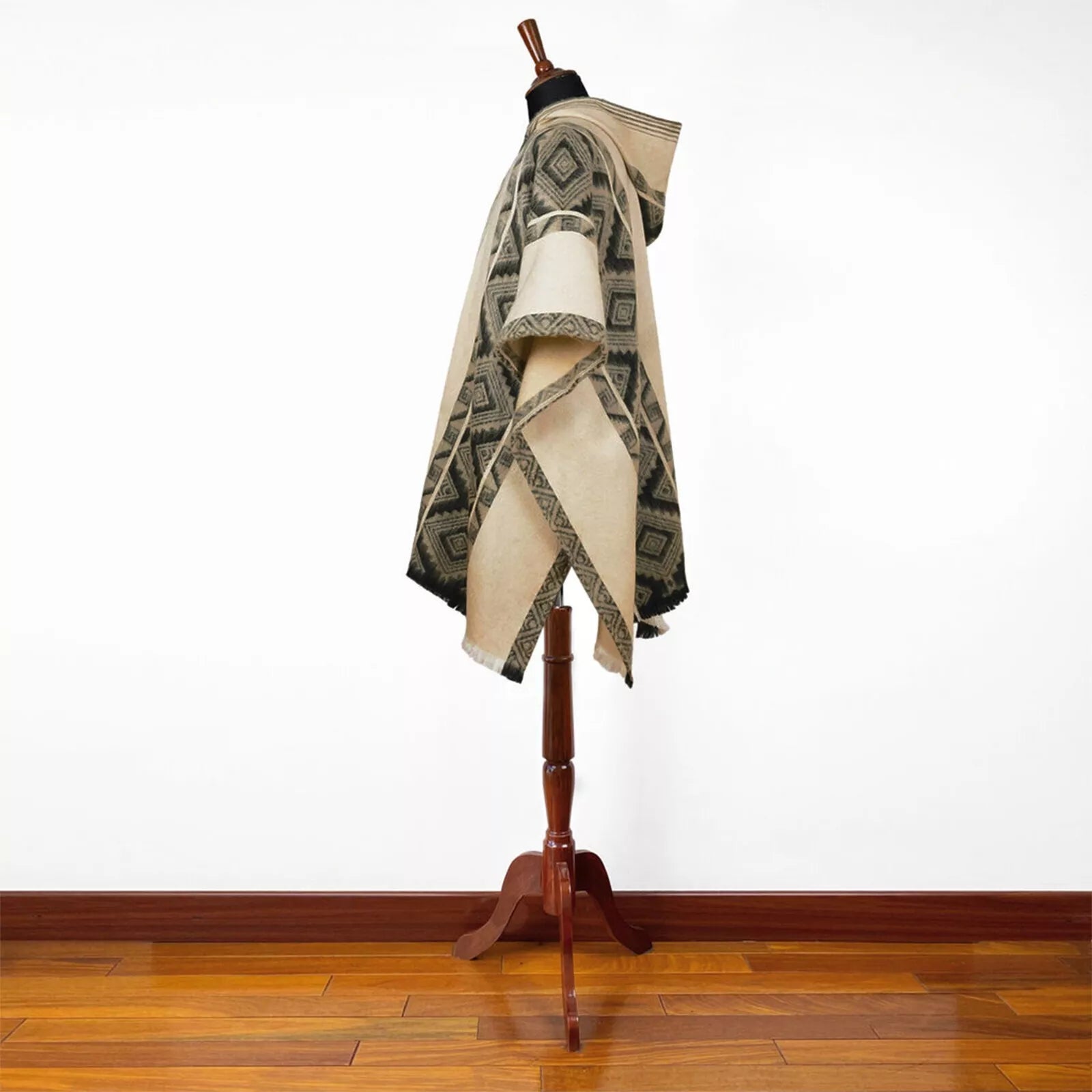 A woven poncho with geometric black patterns drapes over a wooden mannequin stand, set against a white wall and wooden floor background.