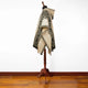 A woven poncho with geometric black patterns drapes over a wooden mannequin stand, set against a white wall and wooden floor background.