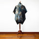 A tie-dye patterned cloak with blue, black, and brown hues hangs on a wooden stand in a room with wooden flooring and a plain white wall.