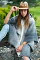 Zuleta llama wool hooded poncho – a person wearing a straw hat and gray poncho sits on a stone ledge. They are outdoors in a garden, surrounded by greenery