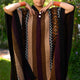A person models a colorful, patterned poncho with geometric designs and stripes. Their hands rest near the neckline. The background shows a lush, green outdoor setting.
