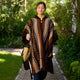 A person wearing a colorful poncho walks along a garden path, holding a woven bag. The lush greenery surrounds them, creating a serene outdoor setting.