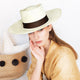 A person wearing a light-colored wide-brimmed hat with a dark band rests their chin on their hand beside a textured basket with decorative black dots, set against a plain background.