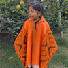 A child wearing an orange poncho with geometric patterns is smiling. They stand in a garden with lush green foliage and visible fruit on the trees.