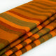 Putumayo baby alpaca wool blanket – a folded woolen blanket displays orange and dark greenish-brown stripes, arranged on a smooth white surface, highlighting its texture and warmth in a simple, uncluttered setting