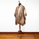 A mannequin displays a beige poncho with geometric patterns and fringe, standing on a wooden floor against a plain white wall.
