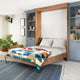 A wooden Murphy bed partially unfolded, features a colorful geometric blanket and gray pillows, positioned between built-in shelves filled with books, plants, and decor in a cozy, carpeted room.