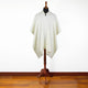 A cream-colored poncho, draped over a mannequin, is standing on a wooden stand against a plain white wall and hardwood floor.