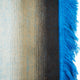 A woven textile displays vertical, gradient stripes transitioning from light blue to brown, with bright blue fringes on the right, sitting against a plain white background.