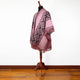 A patterned pink poncho with fringe detail is draped over a wooden mannequin, set in a room with polished wooden flooring and a plain white wall.