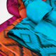 Upano kids alpaca hooded poncho – colorful fabric pieces lie overlapping, displaying vibrant turquoise, orange, and magenta hues with intricate patterns. The scene's layout emphasizes the fabric textures in a static arrangement