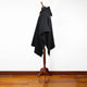 A black poncho with a hood is draped on a wooden mannequin stand, placed against a plain white wall, on a hardwood floor.