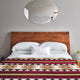 A bed with white and red patterned bedding stands against a gray wall. A wooden headboard supports white pillows. An oval mirror with decorative etching hangs above. A small plant rests on a bedside table.