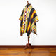 A multicolored poncho with geometric patterns is displayed on a wooden mannequin stand against a plain white background, resting on a polished wooden floor.
