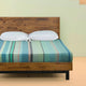 Sucua baby alpaca wool blanket – a wooden bed with a striped green and blue bedspread is topped with two white pillows. It stands against a light-colored wall on a beige floor
