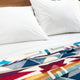 Two white pillows rest on a bed with a white sheet and a colorful patterned blanket featuring red, blue, and yellow geometric designs, all set against a wooden headboard.