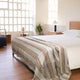 Quijos alpaca wool blanket – a bed is made with white sheets and a striped blanket, placed in a bright room with wooden floors. Nearby, a wooden nightstand holds a lamp and a potted plant
