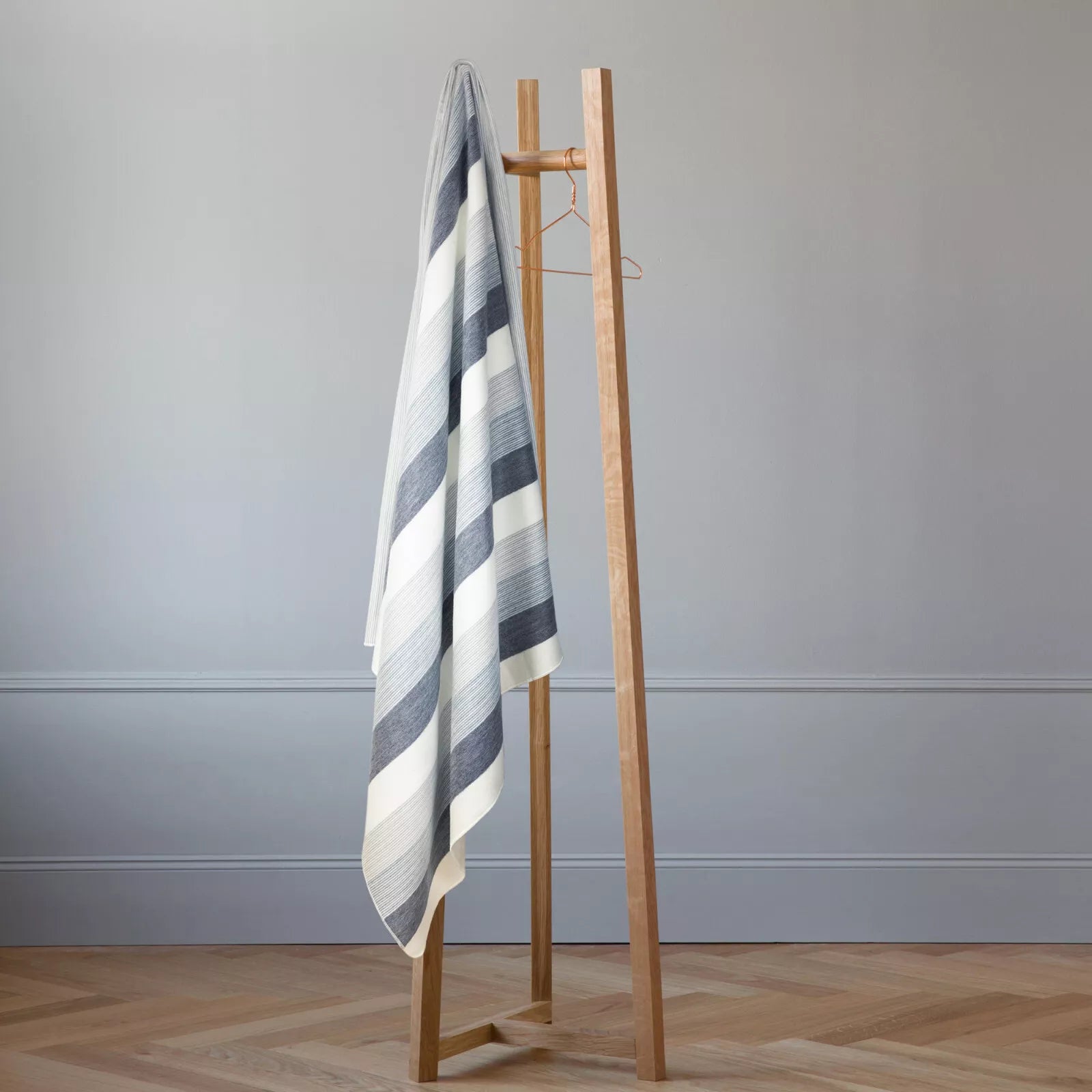 A striped gray and white towel hangs on a simple wooden rack. Two empty hangers are present. The scene is set in a minimalistic room with light-colored walls and parquet flooring.