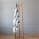 Cayambe baby alpaca wool blanket – a striped gray and white blanket hangs on a simple wooden rack. Two empty hangers are present. The scene is set in a minimalistic room with light-colored walls and parquet flooring