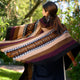 A woman drapes a colorful, geometric-patterned shawl over her shoulders while walking in a lush, green garden, surrounded by bright sunlight and verdant foliage.