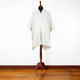 A white poncho with a hood and button closure is displayed on a wooden mannequin stand. The background is a plain white wall, and the floor is hardwood.