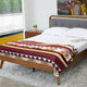 A bed with a colorful patterned blanket lies neatly on a light gray rug. A wooden nightstand with a small potted plant sits nearby, under a large window.