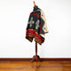 Poncho displayed on a wooden mannequin, featuring intricate geometric patterns in black, red, yellow, and beige. The setting is a simple room with a light-colored wall and wooden floor.