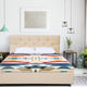 A beige bed with a colorful geometric-patterned blanket sits in a minimalist bedroom, featuring hanging lights, a side table with plants, and a framed artwork on a white wall.