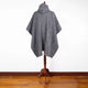 A gray, textured poncho with a hood is displayed on a wooden mannequin stand against a simple white wall backdrop with wooden flooring.