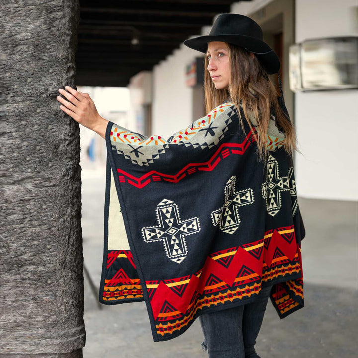 A person wearing a colorful patterned poncho and black hat leans against a tree trunk in an outdoor setting with a white building in the background.