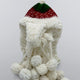 A white, fluffy knit hat with braids and pom-poms rests against a plain background, featuring a green and red patterned top.