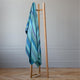 Pululahua baby alpaca wool blanket – a wooden clothes rack holds a striped blue and green blanket draped over it. The scene is set against a plain wall on a wooden parquet floor