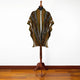 A poncho with geometric patterns in brown, black, and white hangs on a wooden stand. It is displayed in a room with a wooden floor and a plain white wall.