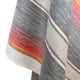 Ajilanga baby alpaca wool blanket – a woven fabric features horizontal stripes in shades of gray, red, orange, and yellow. The cloth hangs draped without visible support, emphasizing its texture and color gradient