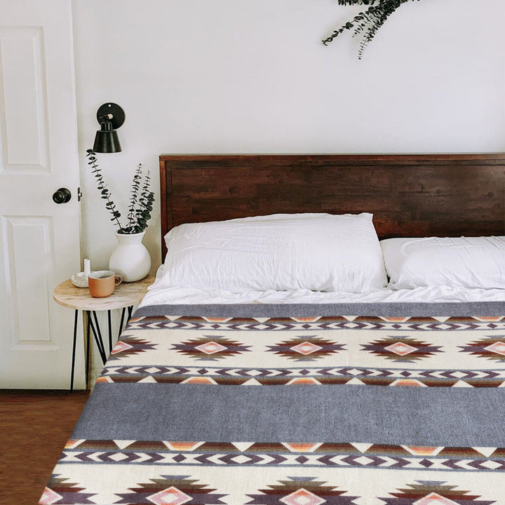 A bed is neatly made with a patterned blanket and white pillows, set against a wooden headboard. A small table beside it holds a potted plant, a mug, and a candle, under a wall sconce.