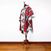 A vibrant poncho hangs on a wooden mannequin stand. It features red, green, and white geometric patterns, situated in a minimalist setting with a wooden floor and a plain white wall.