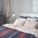 Huasaga baby alpaca wool blanket – bed with colorful striped blanket and light pillows, situated in a softly lit bedroom. A white lamp and dresser with a floral vase add to the serene decor