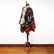 A patterned poncho with geometric designs in red, black, and cream drapes around a mannequin stand, set against a plain white wall and wooden floor background.