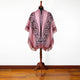 A pink poncho with geometric patterns displayed on a wooden mannequin. The garment features fringed edges and is set against a plain white wall and wooden floor.