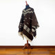 Kids Clint Eastwood alpaca wool poncho – a dark brown poncho with cream geometric patterns hangs on a wooden mannequin. It is displayed in a room with white walls and a polished wooden floor