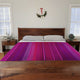 Chone baby alpaca wool blanket – a bed with a vibrant purple and red striped cover is centered in a minimalist bedroom. Two pillows rest against the wooden headboard. Plants and a clock adorn bedside tables