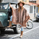 A person wearing a patterned poncho and hat stands beside a rugged, green jeep, parked on a cobblestone street with buildings and a motorcycle in the background.