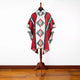A poncho with geometric patterns in red, green, and white is displayed on a wooden stand. It is situated in a room with a wooden floor and white walls.